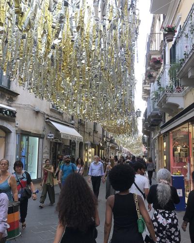 A crowded pedestrian street in an Italian town, lined with shops like "GEOX" and "RUSSOTTI," decorated with hanging gold and silver streamers.