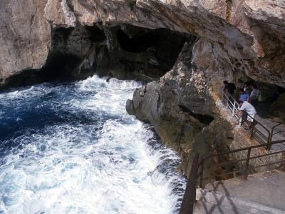 The image shows the interior of a large sea cave with turbulent blue water crashing against rocky walls, and tourists walking along a concrete walkway with metal railings on the right side.
