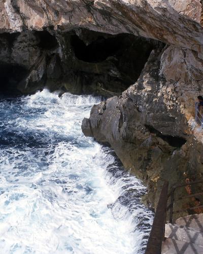 The image shows the interior of a large sea cave with turbulent blue water crashing against rocky walls, and tourists walking along a concrete walkway with metal railings on the right side.