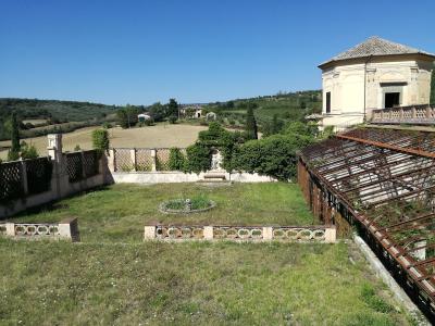 A sunlit garden with a grassy lawn, decorative low walls, a circular flower bed, a trellis structure, and a classical-style building on a hillside overlooking rural Umbrian countryside.