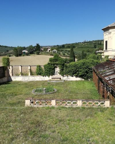 A sunlit garden with a grassy lawn, decorative low walls, a circular flower bed, a trellis structure, and a classical-style building on a hillside overlooking rural Umbrian countryside.