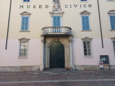 The image shows the facade of a pale yellow and pink building with blue shutters, featuring a central balcony above an arched doorway, with large black letters spelling "MUSEO CIVICO" on the upper wall and a small sign to the right reading "COMUNA