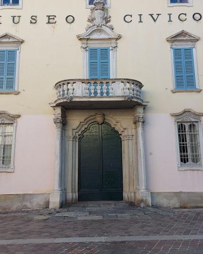 The image shows the facade of a pale yellow and pink building with blue shutters, featuring a central balcony above an arched doorway, with large black letters spelling "MUSEO CIVICO" on the upper wall and a small sign to the right reading "COMUNA