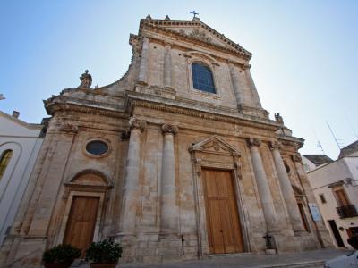 A low-angle photograph of a large, Baroque-style stone church facade featuring a central pediment, pilasters, a large wooden door, and statues on the roofline, set against a clear sky with adjacent buildings.