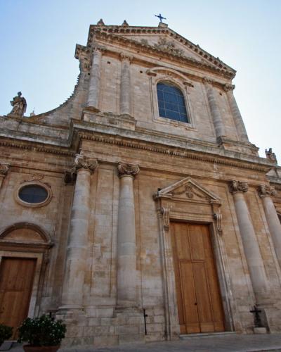 A low-angle photograph of a large, Baroque-style stone church facade featuring a central pediment, pilasters, a large wooden door, and statues on the roofline, set against a clear sky with adjacent buildings.