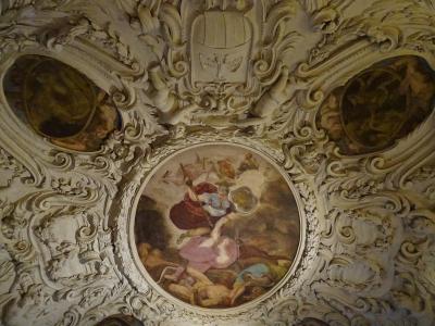An ornate, white stucco ceiling featuring a central circular fresco of mythological figures, flanked by two smaller oval paintings and topped with a coat of arms, identified as the Giunta Albertiana room.