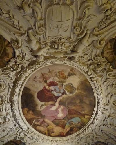 An ornate, white stucco ceiling featuring a central circular fresco of mythological figures, flanked by two smaller oval paintings and topped with a coat of arms, identified as the Giunta Albertiana room.