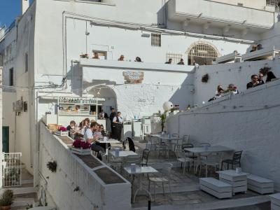 The image shows an outdoor terrace of a restaurant or bistro named "Borgo Antico Bistrot," featuring white-washed buildings, stone steps, and patrons dining under a clear blue sky.