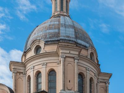 A low-angle photograph of a large Renaissance-style dome with a lantern and cross on top, set against a blue sky with scattered clouds; the structure features arched windows, pilasters, and classical detailing, with adjacent brick buildings visibl