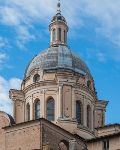 A low-angle photograph of a large Renaissance-style dome with a lantern and cross on top, set against a blue sky with scattered clouds; the structure features arched windows, pilasters, and classical detailing, with adjacent brick buildings visibl