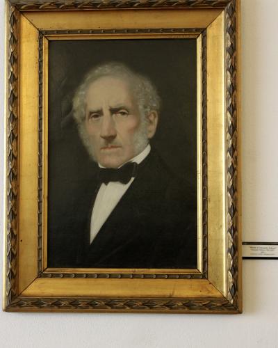 A framed oil portrait of an elderly man with white hair and sideburns, wearing a black suit and bow tie, hanging on a white wall; a small museum label to the right reads “Ritratto di Alessandro Manzoni” by Giuseppe Molteni (1805–1867), dated 1867,