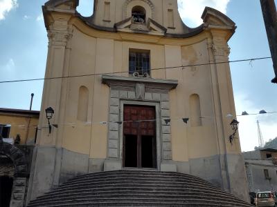 A low-angle photograph of a yellow Baroque-style church facade with a wide, curved stone staircase leading up to a central wooden door, set against a blue sky with clouds.