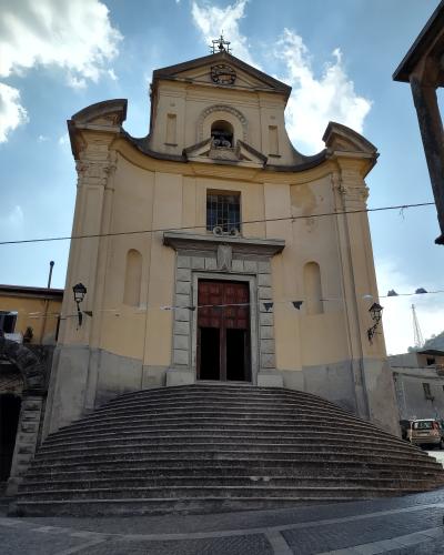 A low-angle photograph of a yellow Baroque-style church facade with a wide, curved stone staircase leading up to a central wooden door, set against a blue sky with clouds.