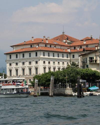 A large white palace with a red-tiled roof situated on a waterfront, with a tour boat in the foreground and a church to the right.