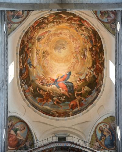 The image shows a large, circular ceiling fresco depicting the Assumption of Mary surrounded by angels and saints, located inside a church with architectural features like arches, windows, and a staircase leading up to the dome; the artwork is sig