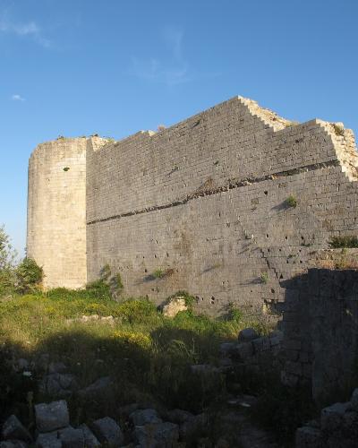 The image shows the stone ruins of a medieval fortress or castle with a cylindrical tower and high walls, set against a blue sky with vegetation in the foreground.