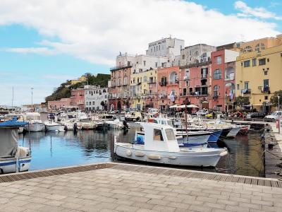 A sunlit harbor scene featuring a row of colorful, multi-story buildings lining a stone-paved waterfront promenade, with numerous small fishing boats and yachts moored in calm blue water under a partly cloudy sky; visible signage includes "ISTITUT