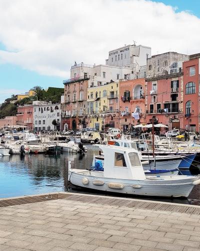 A sunlit harbor scene featuring a row of colorful, multi-story buildings lining a stone-paved waterfront promenade, with numerous small fishing boats and yachts moored in calm blue water under a partly cloudy sky; visible signage includes "ISTITUT
