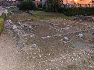 The image shows the excavated archaeological ruins of a Roman site, featuring low stone foundation walls, column bases, and a grassy courtyard, with a modern building and people visible in the background.