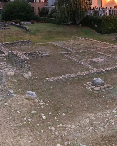 The image shows the excavated archaeological ruins of a Roman site, featuring low stone foundation walls, column bases, and a grassy courtyard, with a modern building and people visible in the background.