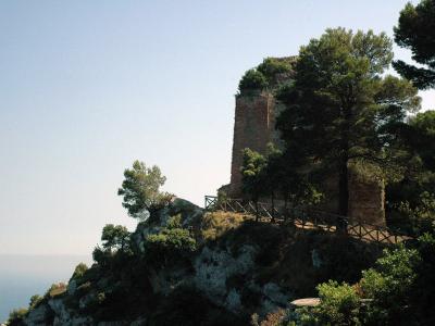 The image shows the ruins of a tall, rectangular brick tower structure perched on a rocky cliff edge, surrounded by Mediterranean pine trees and vegetation, with a wooden safety railing along a path in the foreground and the sea visible in the dis