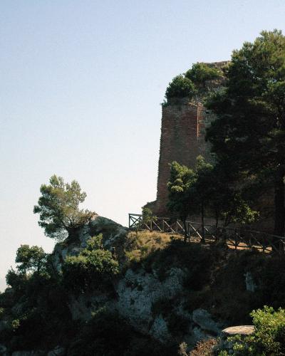 The image shows the ruins of a tall, rectangular brick tower structure perched on a rocky cliff edge, surrounded by Mediterranean pine trees and vegetation, with a wooden safety railing along a path in the foreground and the sea visible in the dis