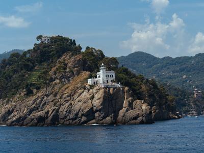 A white lighthouse building with a lantern room sits atop a rocky, tree-covered promontory overlooking the sea, with hills and other buildings visible in the background.