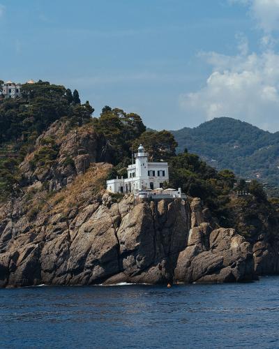 A white lighthouse building with a lantern room sits atop a rocky, tree-covered promontory overlooking the sea, with hills and other buildings visible in the background.