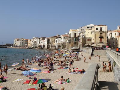 A crowded sandy beach with numerous sunbathers and swimmers, backed by a dense row of multi-story Mediterranean-style buildings with terracotta roofs, under a clear blue sky; a concrete seawall and promenade are visible on the right.