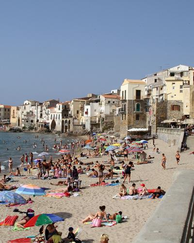 A crowded sandy beach with numerous sunbathers and swimmers, backed by a dense row of multi-story Mediterranean-style buildings with terracotta roofs, under a clear blue sky; a concrete seawall and promenade are visible on the right.
