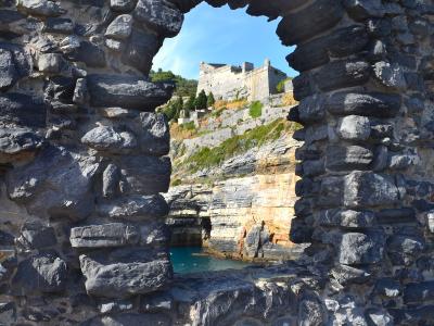 A view through a rough, irregular stone archway in the foreground looking out onto a fortified castle structure perched on a rocky cliffside above turquoise water.