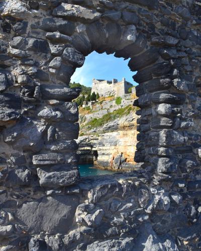 A view through a rough, irregular stone archway in the foreground looking out onto a fortified castle structure perched on a rocky cliffside above turquoise water.