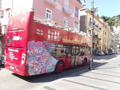 A red double-decker "City Sightseeing Sorrento" open-top tour bus is parked on a paved street lined with multi-story buildings featuring balconies and shuttered windows, under bright daylight.