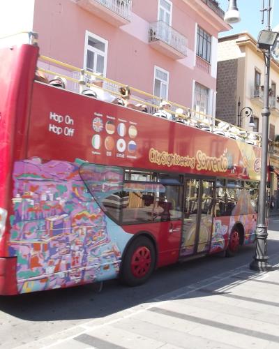 A red double-decker "City Sightseeing Sorrento" open-top tour bus is parked on a paved street lined with multi-story buildings featuring balconies and shuttered windows, under bright daylight.