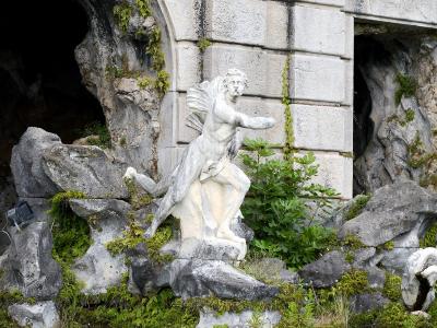 A weathered stone statue of a winged male figure (likely Aeolus) set within a rocky grotto structure with moss and greenery, featuring a modern black spotlight fixture attached to the rocks on the left.