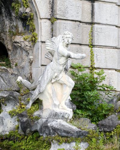 A weathered stone statue of a winged male figure (likely Aeolus) set within a rocky grotto structure with moss and greenery, featuring a modern black spotlight fixture attached to the rocks on the left.