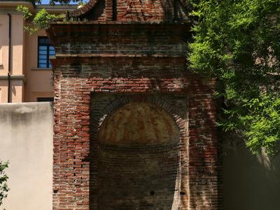 A weathered red-brick architectural niche or small chapel-like structure with an arched recess and decorative top, set within a green garden environment with trees and grass, adjacent to a pale building with a blue-shuttered window.