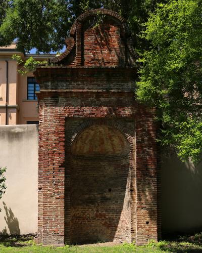 A weathered red-brick architectural niche or small chapel-like structure with an arched recess and decorative top, set within a green garden environment with trees and grass, adjacent to a pale building with a blue-shuttered window.