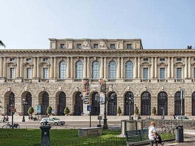 A wide-angle photograph of a large, light-colored neoclassical building with a rusticated ground floor featuring a long arcade of arches and an upper floor with tall arched windows and pilasters, situated in a public square with a park in the fore