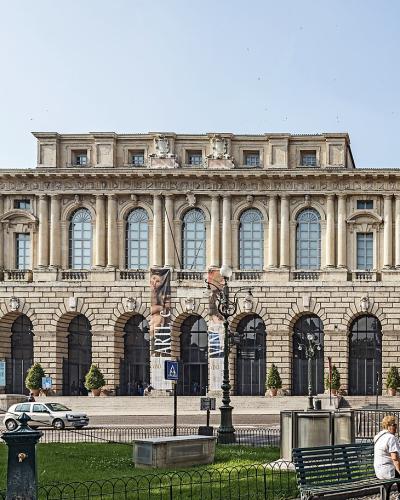 A wide-angle photograph of a large, light-colored neoclassical building with a rusticated ground floor featuring a long arcade of arches and an upper floor with tall arched windows and pilasters, situated in a public square with a park in the fore