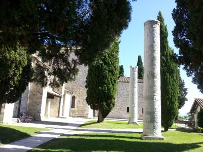 The image shows two tall, weathered stone columns standing on a grassy lawn in front of a large, light-colored stone building with arched windows and a tiled roof, framed by tall cypress trees under a clear blue sky; a bicycle is parked near the b