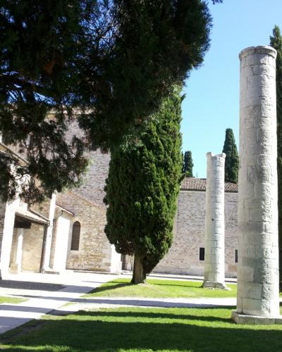 The image shows two tall, weathered stone columns standing on a grassy lawn in front of a large, light-colored stone building with arched windows and a tiled roof, framed by tall cypress trees under a clear blue sky; a bicycle is parked near the b