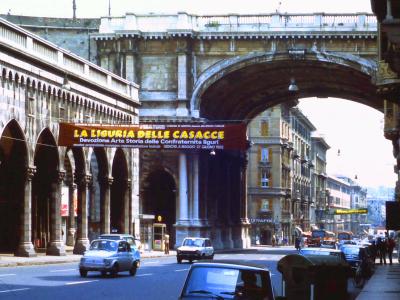 A street scene in Genoa, Italy, featuring the distinctive stone arches of the Acquedotto della Maddalena (Maddalena Aqueduct) spanning over Via XX Settembre, with a large banner advertising an exhibition titled "LA LIGURIA DELLE CASACCE" dated 198