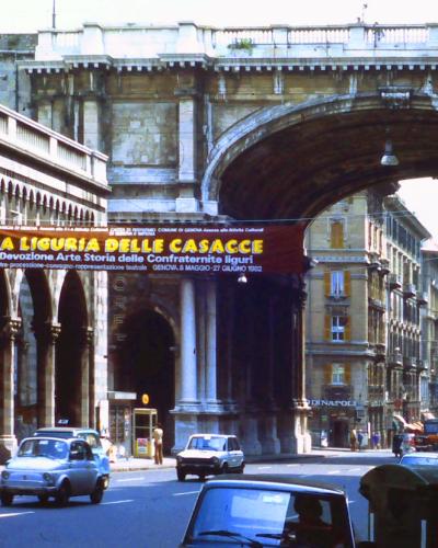 A street scene in Genoa, Italy, featuring the distinctive stone arches of the Acquedotto della Maddalena (Maddalena Aqueduct) spanning over Via XX Settembre, with a large banner advertising an exhibition titled "LA LIGURIA DELLE CASACCE" dated 198