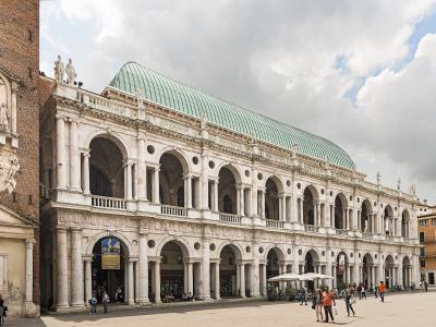 A wide-angle view of the Basilica Palladiana in Vicenza, Italy, showing its distinctive white marble loggias with arched openings on two levels, topped by a green copper roof, situated on Piazza dei Signori with people walking in the foreground an