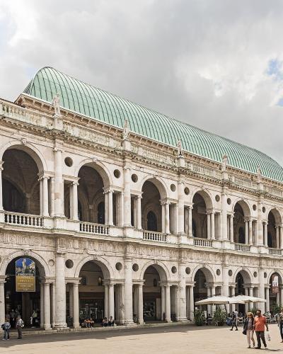 A wide-angle view of the Basilica Palladiana in Vicenza, Italy, showing its distinctive white marble loggias with arched openings on two levels, topped by a green copper roof, situated on Piazza dei Signori with people walking in the foreground an