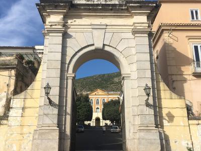 The image shows a large, ornate stone archway with a royal coat of arms and lion statues on top, leading into a courtyard with a yellow building in the background under a blue sky.