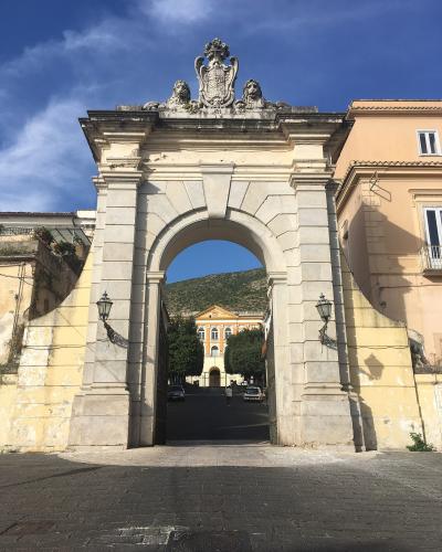 The image shows a large, ornate stone archway with a royal coat of arms and lion statues on top, leading into a courtyard with a yellow building in the background under a blue sky.