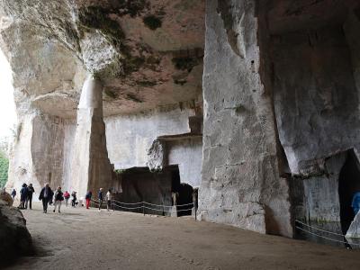 A view of a limestone cliff face with large natural and man-made caves, including a prominent arched entrance on the left, surrounded by dense Mediterranean vegetation and trees under an overcast sky.