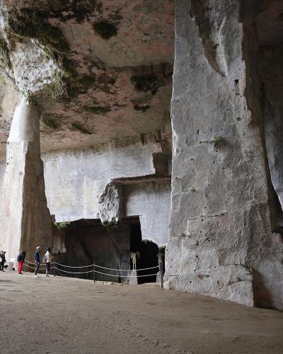 A view of a limestone cliff face with large natural and man-made caves, including a prominent arched entrance on the left, surrounded by dense Mediterranean vegetation and trees under an overcast sky.